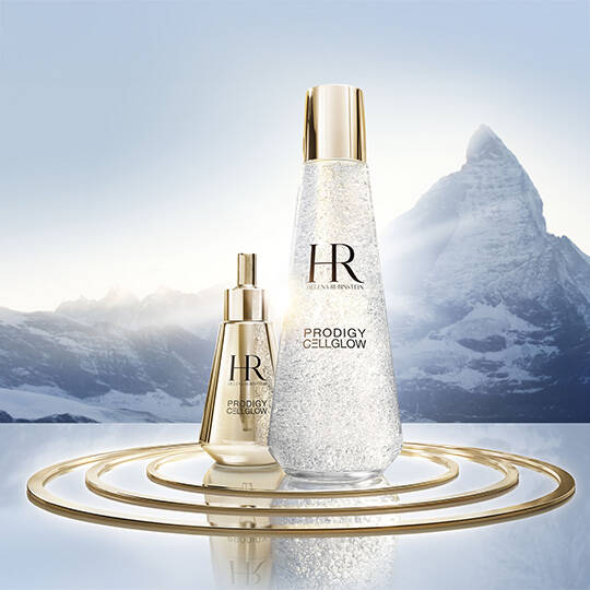 Helena Rubinstein Prodigy Cellglow Dewy Essence and Oilixir are displayed on gold rings against a backdrop of a snowy mountain reflected in a lake.​